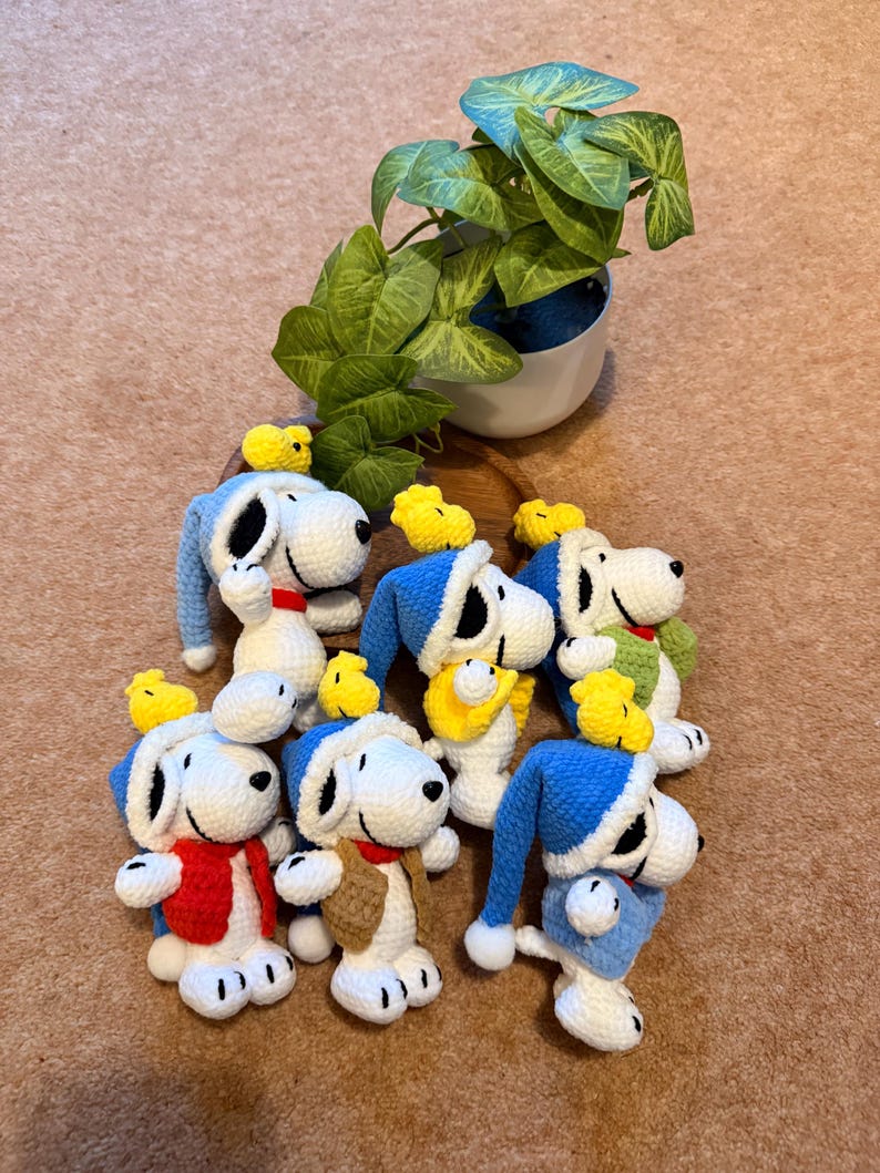 May include: A collection of handmade crochet Snoopy figures, each with a unique outfit and a small yellow bird. The Snoopy figures are primarily white with black accents, wearing blue, red, and brown clothing. A potted plant is in the background.