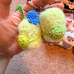 May include: A pair of handmade, fuzzy keychains in the shape of birds. One keychain is yellow and green, with a blue accent. The other is yellow and green. Both are attached to a metal keyring with a green chain.