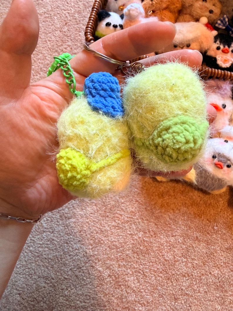 May include: A pair of handmade, fuzzy keychains in the shape of birds. One keychain is yellow and green, with a blue accent. The other is yellow and green. Both are attached to a metal keyring with a green chain.