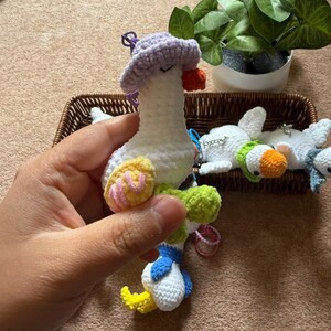 Puede incluir: Un ganso de ganchillo hecho a mano con cuerpo blanco, sombrero lila y detalles coloridos. El ganso tiene pico y patas amarillas, y alas azules y blancas. Otros gansos de ganchillo est&aacute;n en una cesta de mimbre.