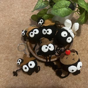 May include: A collection of handmade crochet keychains featuring black, round characters with large white eyes. Some characters have brown hats or accessories, and one has white bunny ears. The keychains are displayed on a wooden plate.