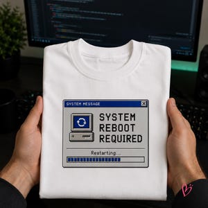 May include: A white t-shirt featuring a graphic of a retro computer system message window. The text reads SYSTEM MESSAGE and SYSTEM REBOOT REQUIRED, with a pixelated computer icon and a progress bar labeled Restarting below the main text.