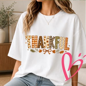 Op de afbeelding: Een wit T-shirt met korte mouwen en het woord THANKFUL in grote blokletters. Elke letter heeft een ander patroon, waaronder luipaardprint, ruitjes en herfstbladeren. Onder de tekst staan kleine bladillustraties en een strik afgebeeld.