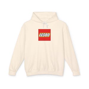 Pode incluir: Uma camisola com capuz de cor creme com um gráfico quadrado vermelho no peito. Dentro do quadrado vermelho está o texto LESBO escrito em letras brancas com um contorno preto grosso e uma borda amarela fina, estilo logo de bloco.
