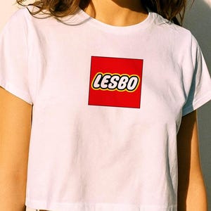 May include: White crop top with a red square graphic that says "LESBO" in yellow and white block letters. The shirt is a casual style, suitable for everyday wear.