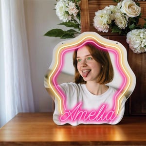 May include: A white neon sign with a pink neon outline in the shape of a star. The sign has a photo of a person with red hair and green eyes in the center. The name "Amelia" is written in pink neon letters at the bottom of the sign.