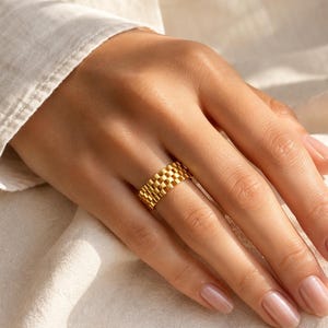 Könnte beinhalten: Ein goldfarbener Ring mit einem sich wiederholenden Gliedermuster, das an die Struktur eines Uhrenarmbands erinnert. Der Ring hat eine polierte, metallische Oberfläche mit einem durchgehenden, strukturierten Design auf der Außenseite.