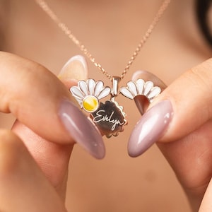Könnte beinhalten: Eine roségoldene Halskette mit einem blumenförmigen Medaillon-Anhänger mit weißen Blütenblättern und gelber Mitte. Das Medaillon lässt sich öffnen und zeigt einen runden Anhänger, der mit dem Namen Evelyn in Schreibschrift graviert ist.