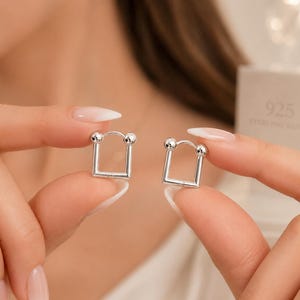 Può includere: Un paio di orecchini a cerchio in argento con un design quadrato e un piccolo dettaglio a sfera su ogni angolo superiore. Gli orecchini sono realizzati in argento sterling, come indicato dal marchio 925 visibile sulla confezione sullo sfondo.