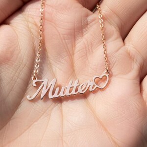 May include: A rose gold necklace with the word "Mutter" in cursive script, ending with a heart-shaped charm. The delicate chain is visible against a hand, showcasing the jewelry's design and color.
