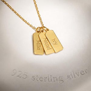 Può includere: Una collana color oro con tre piccoli ciondoli rettangolari appesi a una catenina sottile. Ogni targhetta è incisa con un nome: Mila, Hazel e Ivy. La collana è esposta su un oggetto bianco, liscio e rotondo con la scritta 925 sterling silver.