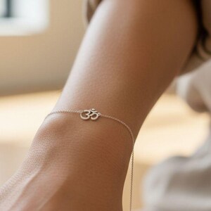 May include: A delicate silver bracelet featuring the Om symbol. The bracelet is a fine chain with the Om symbol centered. The bracelet is worn on a wrist, with the arm in a neutral position. The background is blurred.