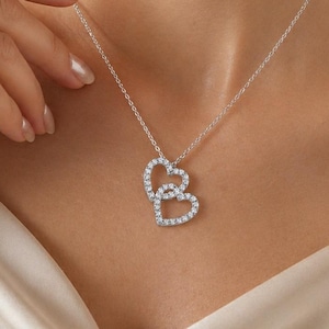 Peut inclure: Un collier en argent avec un pendentif en forme de deux cœurs entrelacés, chacun orné de petites pierres claires et scintillantes. Le collier est porté sur un haut crème, la chaîne reposant sur la peau.