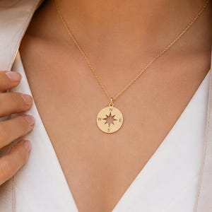 May include: A gold-colored compass pendant necklace. The pendant is round with a star design in the center and directional markings. The necklace is worn by a person wearing a white top and a light-colored jacket.