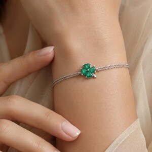 May include: A silver bracelet featuring a four-leaf clover charm with vibrant green gemstones. The clover is centered on a delicate chain, creating a simple yet elegant design. The bracelet is shown on a wrist.