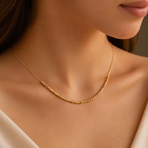 May include: A gold-toned necklace featuring a delicate chain adorned with a series of small, cylindrical beads and tiny spherical beads arranged in a repeating pattern along the centre section, resting against the collarbone area.