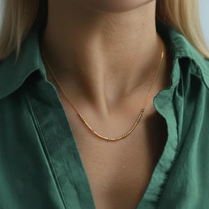 May include: A delicate gold necklace with a series of small, evenly spaced beads and bars. The necklace is displayed against a green collared shirt. The necklace is a simple, elegant piece of jewelry.