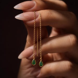 Pode incluir: Um par de brincos de fio de ouro com correntes finas e delicadas e pedras preciosas verdes em forma de gota. Os brincos são projetados para serem passados pela orelha, com as correntes douradas penduradas para exibir as pedras verdes.
