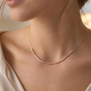 May include: A rose gold-toned necklace featuring a delicate chain adorned with a series of small, cylindrical and spherical metal beads arranged in a sequence along the center, creating a subtle, linear pattern across the collarbone area.