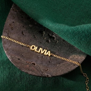 Op de afbeelding: Een goudkleurige kettingarmband met de naam "OLIVIA" in blokletters. De armband ligt op een donker, getextureerd oppervlak tegen een groene achtergrond.