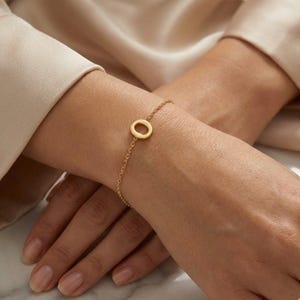 Könnte beinhalten: Ein zartes goldenes Armband mit einem kleinen, kreisförmigen Anhänger. Das Armband ist eine feine Kette, und der Anhänger ist ein einfacher, offener Kreis. Der Schmuck wird am Handgelenk getragen, und die Gesamtästhetik ist minimalistisch und elegant.