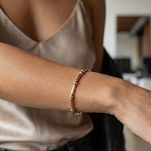 May include: A gold bracelet with a series of small, round beads and elongated cylindrical segments. The bracelet is worn on a person's arm, with a neutral background.
