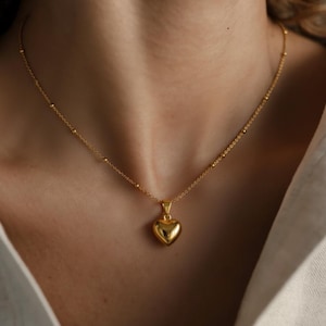 May include: A gold-colored necklace with a heart-shaped pendant. The necklace features a delicate chain with small, evenly spaced beads. The heart pendant is smooth and rounded, reflecting light. The necklace is worn against a neutral background.