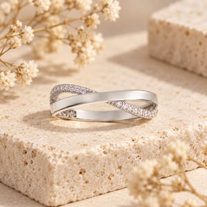 May include: A silver-toned ring featuring a crossover design where one band is smooth and polished, while the other is encrusted with small, clear, sparkling stones. The ring has a continuous, intertwined structure that creates a balanced look.