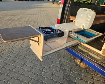May include: A wooden countertop with a fold-down table, a small gas stove, a pull-out drawer with a sink, and a water jug. The countertop is mounted in the back of a van.