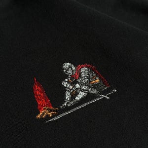 Pode incluir: Uma t-shirt preta com um bordado detalhado de um cavaleiro em armadura prateada com uma capa vermelha, sentado ao lado de uma pequena fogueira de cores vermelho e laranja vibrantes. Uma espada longa repousa no chão junto ao cavaleiro.