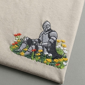 Könnte beinhalten: Gesticktes Design eines Ritters in silberner Rüstung, der sich in einem Feld bunter Blumen ausruht. Der Ritter sitzt in einem Fleck grünen Grases mit gelben, roten und weißen Blumen. Der Hintergrund ist ein beiger Stoff.