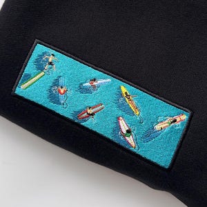 close-up embroidered surf scene patch with surfers on colorful boards in blue ocean water on black hoodie, summer beach aesthetic, surf vibes clothing