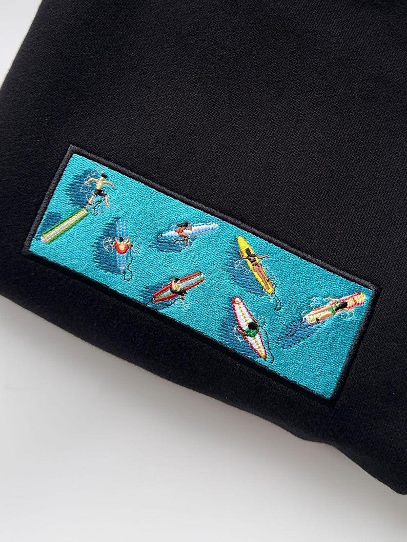 close-up embroidered surf scene patch with surfers on colorful boards in blue ocean water on black hoodie, summer beach aesthetic, surf vibes clothing