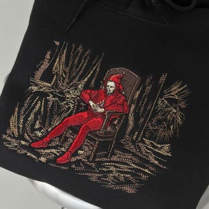 Peut inclure: Un sweat-shirt à capuche noir orné d'une broderie complexe représentant un personnage en tenue de bouffon rouge assis sur une chaise. La broderie utilise des fils rouges, bruns et dorés pour illustrer le personnage et le feuillage environnant.