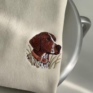 May include: A beige sweatshirt featuring a detailed embroidery of a brown and white hunting dog head. The dog is depicted with a brown collar, surrounded by embroidered green grass blades, positioned on the lower right side of the garment.