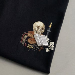 Könnte beinhalten: Ein schwarzes Kleidungsstück aus Stoff mit einer detaillierten Stickerei, die einen menschlichen Schädel auf zwei braunen Büchern, ein offenes Buch, eine brennende Kerze in einem dunklen Halter, ein kleines Holzkreuz und weiße Blüten zeigt.