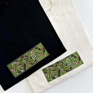 Pu&ograve; includere: Due magliette, una nera e una bianco sporco, con una toppa rettangolare ricamata. La toppa presenta uno sfondo verde con un motivo di pesci koi rossi, neri e bianchi, in un design a vortice.