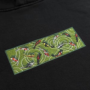 Pu&ograve; includere: Felpa con cappuccio nera con una toppa ricamata raffigurante carpe koi in uno stagno. La toppa ha uno sfondo verde con disegni di increspature bianche. Le carpe koi sono in vari colori, tra cui rosso, arancione, giallo e nero.