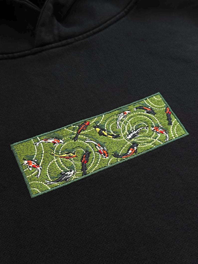 Pu&ograve; includere: Felpa con cappuccio nera con una toppa ricamata raffigurante carpe koi in uno stagno. La toppa ha uno sfondo verde con disegni di increspature bianche. Le carpe koi sono in vari colori, tra cui rosso, arancione, giallo e nero.