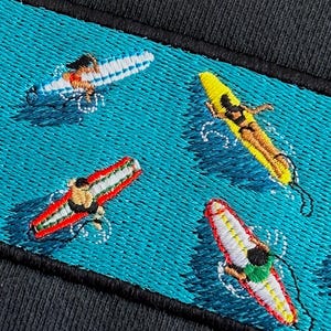 Peut inclure: Broderie sur un tissu bleu fonc&eacute; repr&eacute;sentant quatre personnes faisant du surf sur des planches de surf de diff&eacute;rentes couleurs. L'arri&egrave;re-plan est de couleur bleu turquoise.