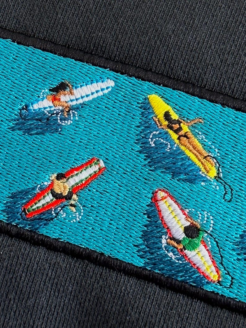 Peut inclure: Broderie sur un tissu bleu fonc&eacute; repr&eacute;sentant quatre personnes faisant du surf sur des planches de surf de diff&eacute;rentes couleurs. L'arri&egrave;re-plan est de couleur bleu turquoise.