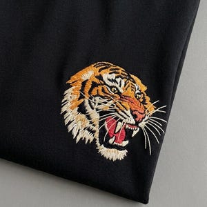 Pode incluir: Uma peça de tecido preto com uma cabeça de tigre bordada detalhada. O tigre é retratado com pelo laranja, preto e branco, nariz rosa e boca aberta mostrando dentes brancos e língua vermelha, com longos bigodes brancos que se estendem para fora.