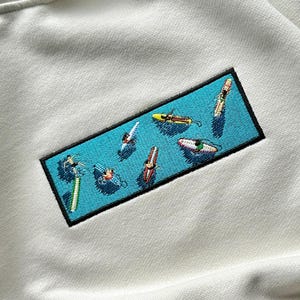 Peut inclure: Un sweat-shirt blanc avec un patch brod&eacute; bleu et noir repr&eacute;sentant une sc&egrave;ne de personnes faisant du surf sur des vagues. Le patch a un fond bleu clair avec des contours blancs et les personnes sont repr&eacute;sent&eacute;es dans diff&eacute;rentes couleurs.