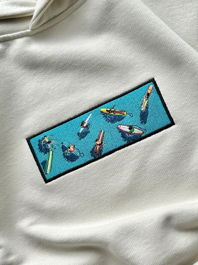 Peut inclure: Un sweat-shirt blanc avec un patch brod&eacute; bleu et noir repr&eacute;sentant une sc&egrave;ne de personnes faisant du surf sur des vagues. Le patch a un fond bleu clair avec des contours blancs et les personnes sont repr&eacute;sent&eacute;es dans diff&eacute;rentes couleurs.