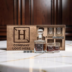 Può includere: Un set in scatola di legno con una caraffa, due bicchieri e pietre per whisky. La caraffa e i bicchieri presentano un logo con la lettera "H" e il testo "COMPANY LOGO HERE". La scatola mostra anche lo stesso logo e testo.