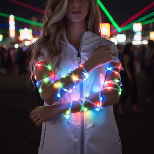 Op de afbeelding: Een witte jas met ritssluiting met veelkleurige LED-lampjes om de armen gewikkeld. De lampjes zijn rood, groen, blauw en geel. De achtergrond is vervaagd met andere lichten.