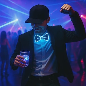 Op de afbeelding: Een persoon draagt een zwarte blazer, een zwarte pet en een blauwe, oplichtende vlinderdas. De persoon houdt een glas water met ijs vast. De achtergrond is een drukke dansvloer met blauwe en paarse verlichting.