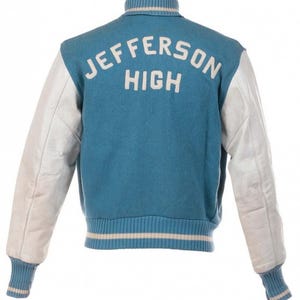 Puede incluir: Chaqueta universitaria azul claro con mangas de cuero blanco. La parte trasera de la chaqueta muestra el texto "JEFFERSON HIGH" en blanco. Cuello, puños y cintura de canalé con rayas blancas. Prenda de vestir clásica.