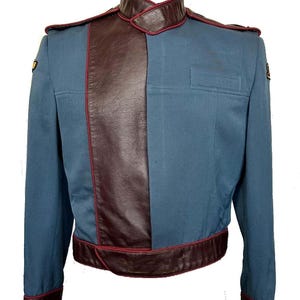 May include: A blue and burgundy jacket with a leather panel down the front. The jacket has a high collar and a belt. There are two gold badges on the jacket.