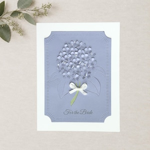 May include: A rectangular greeting card with a white border and a periwinkle blue center panel. The center features a die-cut hydrangea flower design with a small white ribbon bow at the stem and the text For the Bride printed in dark gray.